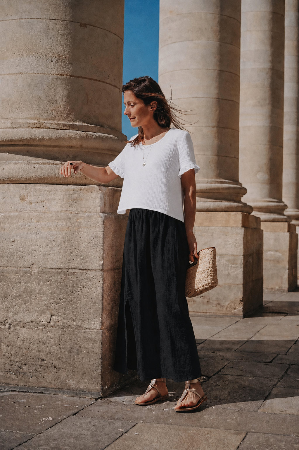 Silhouette complète : jupe longue Lison noire, top blanc et panier en osier, sous les colonnes